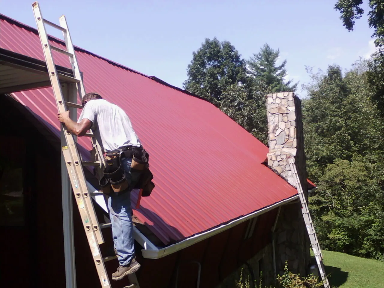 Roofer on a ladder beside a red metal roof and stone chimney — Soffit & Fascia Repair by Roofing Brothers in Oak Hall